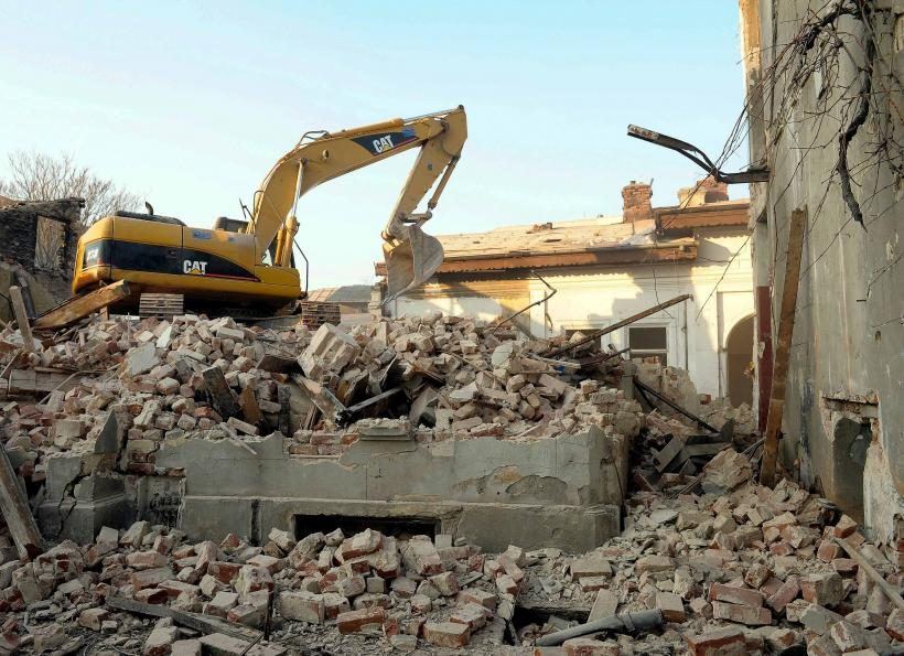 CAT kotrógép téglatörmelék-halmon, részben lebontott régi téglaépület és betonpince-falak