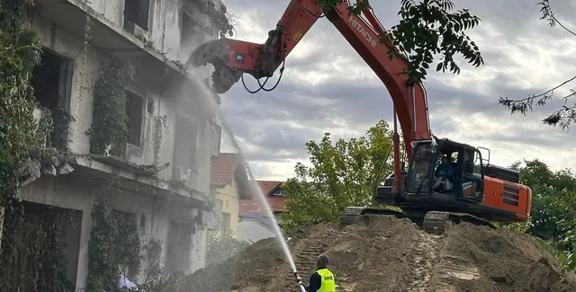 Hitachi lánctalpas kotró törőollóval bont egy elhagyott, többszintes vasbeton épületet, előtérben vízsugaras porkötés