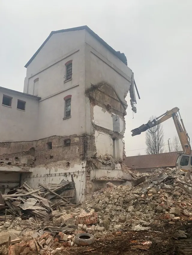 Négyszintes régi ipari épület felülről lefelé történő bontása Liebherr kotró hosszú gémjére szerelt bontóollóval