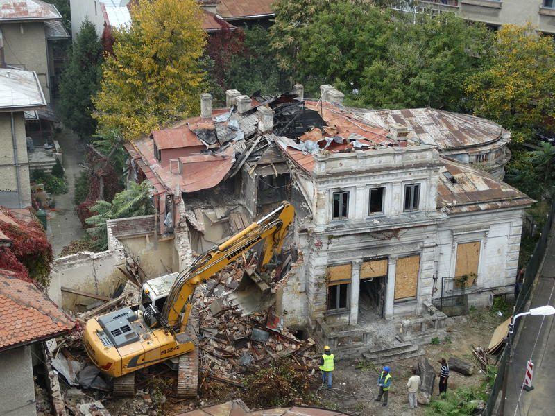 Felülnézeti kép: Liebherr kotró bont egy régi, tetőkupolás villaépületet sűrűn beépített városi környezetben, láthatósági mellényes munkatársakkal